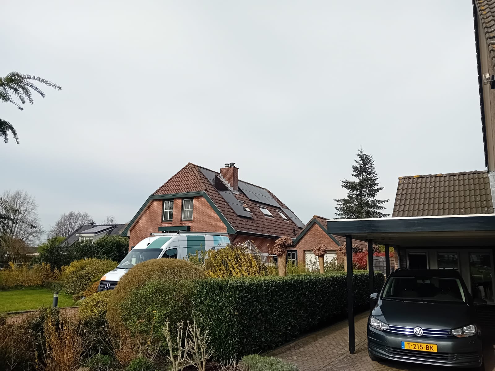 De foto toont een woning met zonnepanelen op het dak, omgeven door tuin en voertuigen.