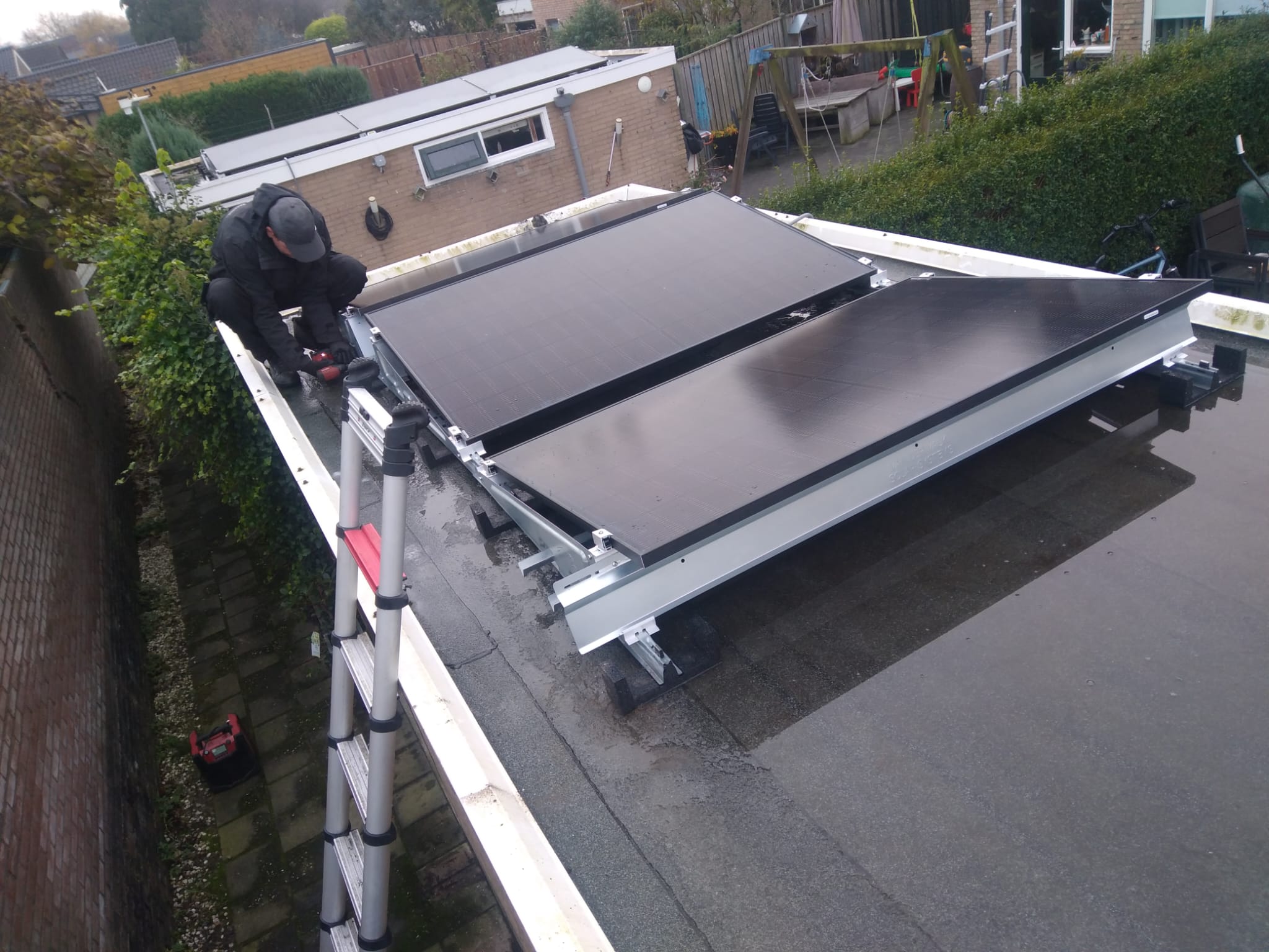 De afbeelding toont een technicus die zonnepanelen installeert op een plat dak, met een ladder voor ondersteuning.