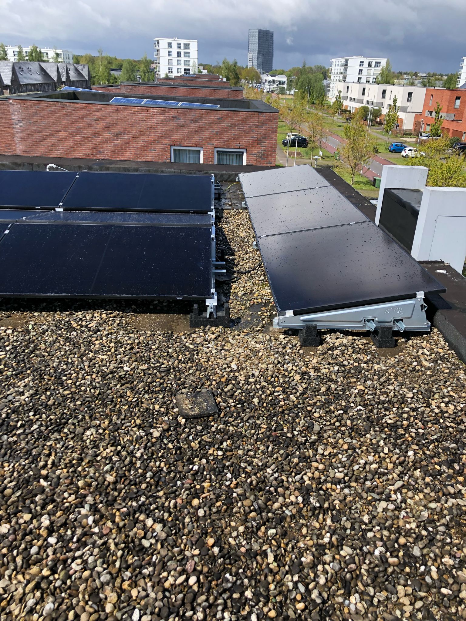 De afbeelding toont een reeks zonnepanelen op een plat dak, met een zichtbaar grindbed en omliggende gebouwen op de achtergrond.