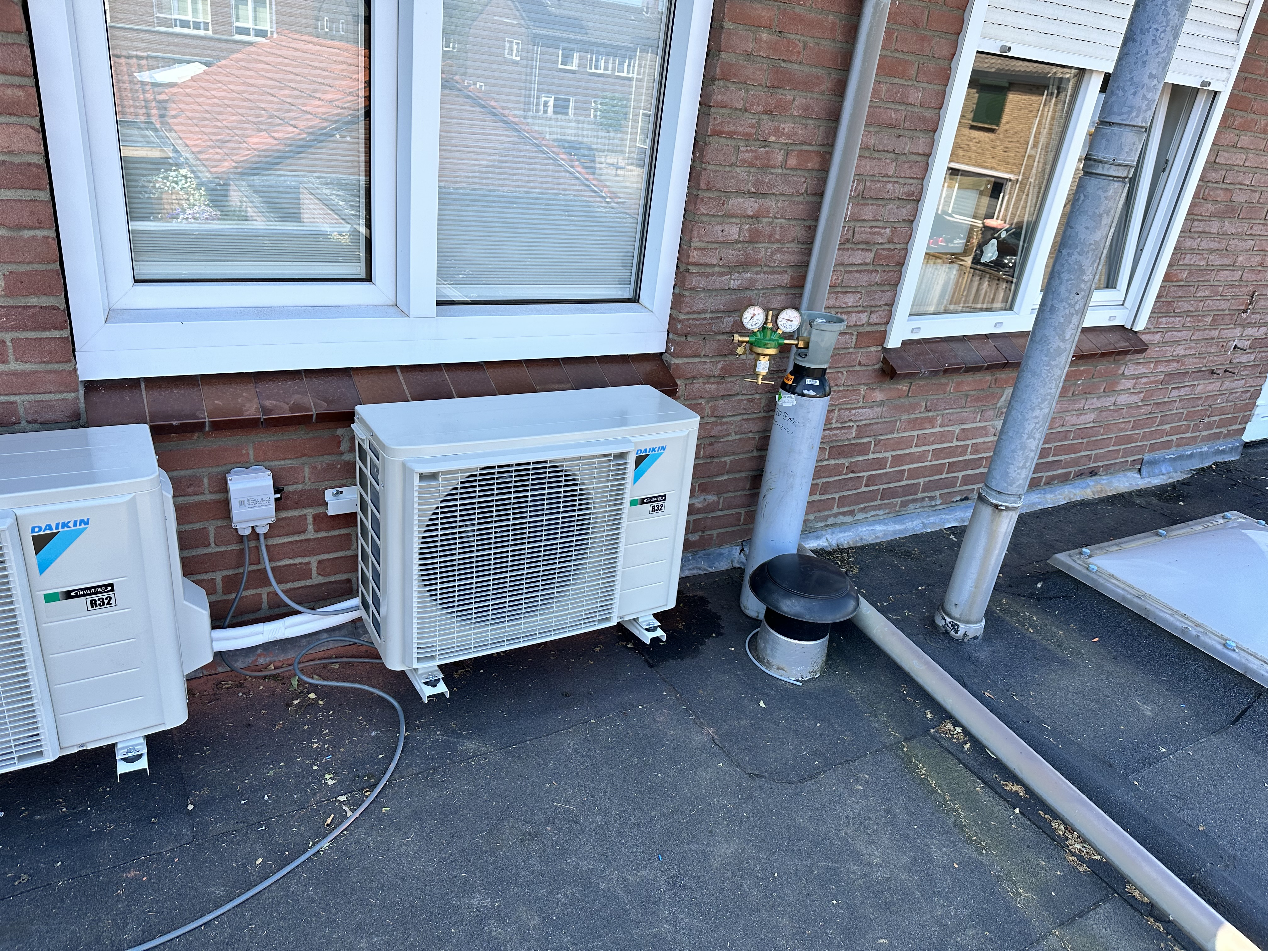 De afbeelding toont een buitenunit van een airconditioninginstallatie. Er zijn twee units zichtbaar, met bijbehorende leidingen en aansluitingen.