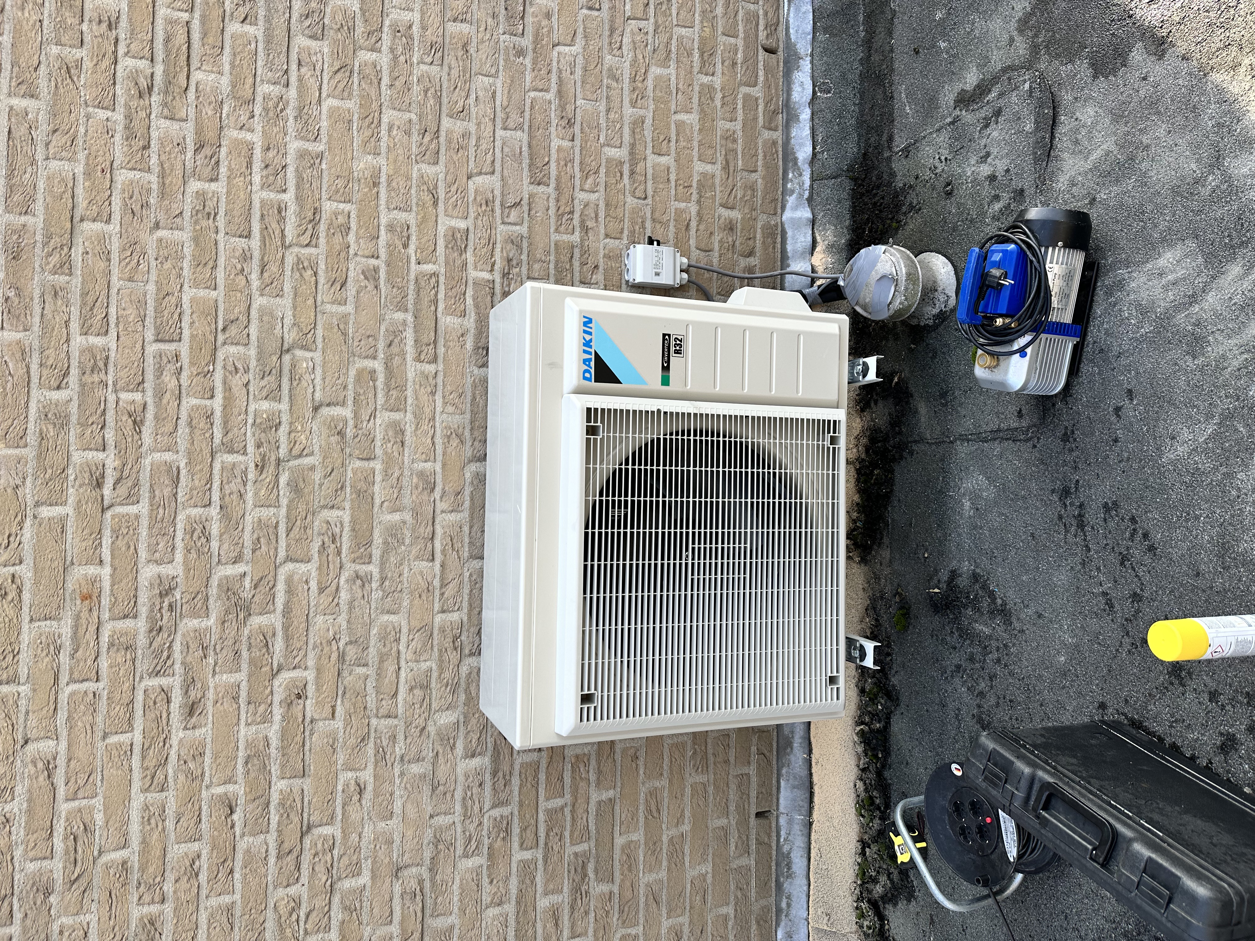De afbeelding toont een buitenunit van een Daikin airconditioning systeem, geïnstalleerd tegen een bakstenen muur, met verschillende gereedschappen en accessoires op de vloer.
