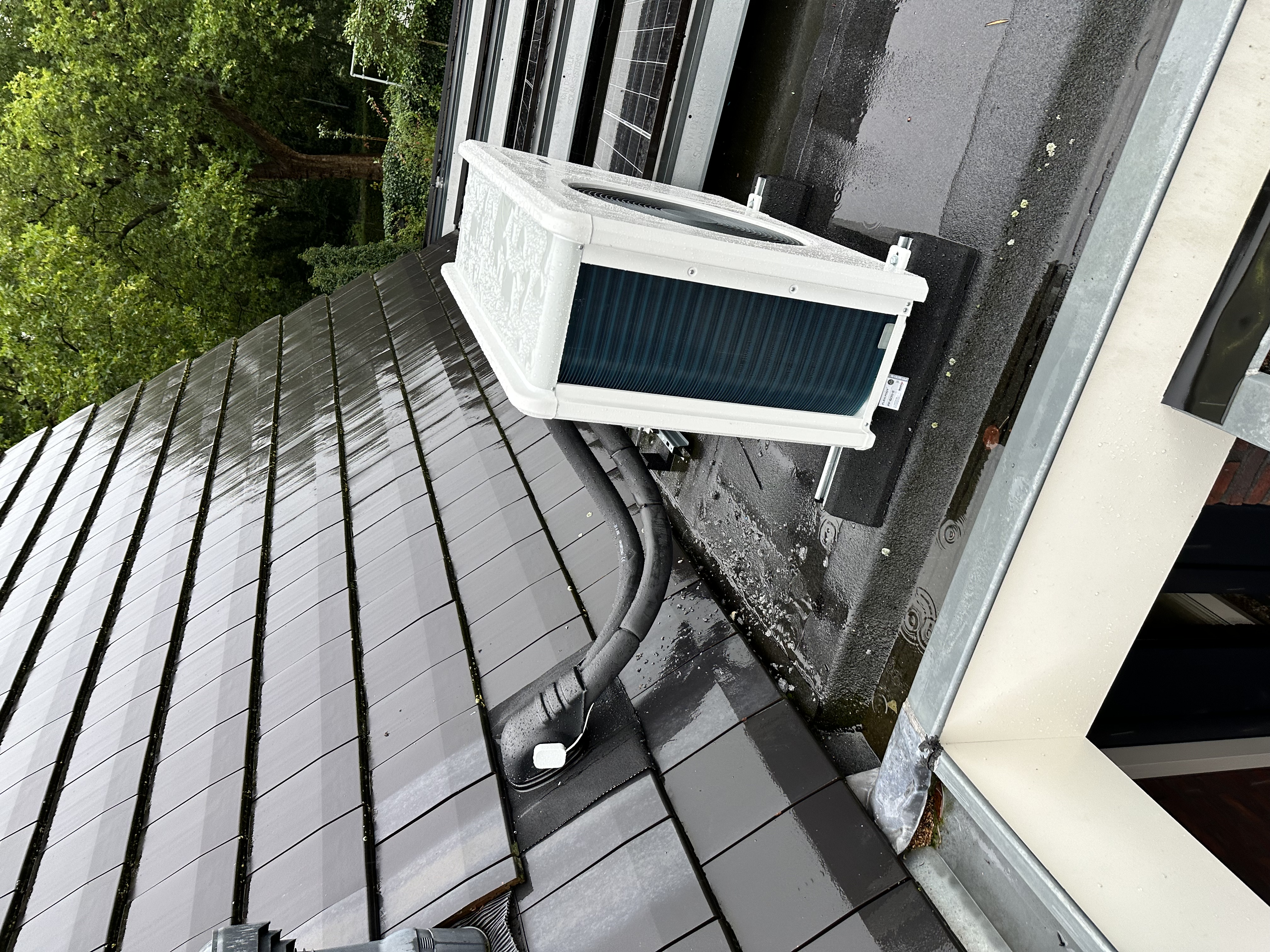 Installatie van een warmtepomp op een dak, met zichtbare aansluitingen en zonnepanelen op de achtergrond.