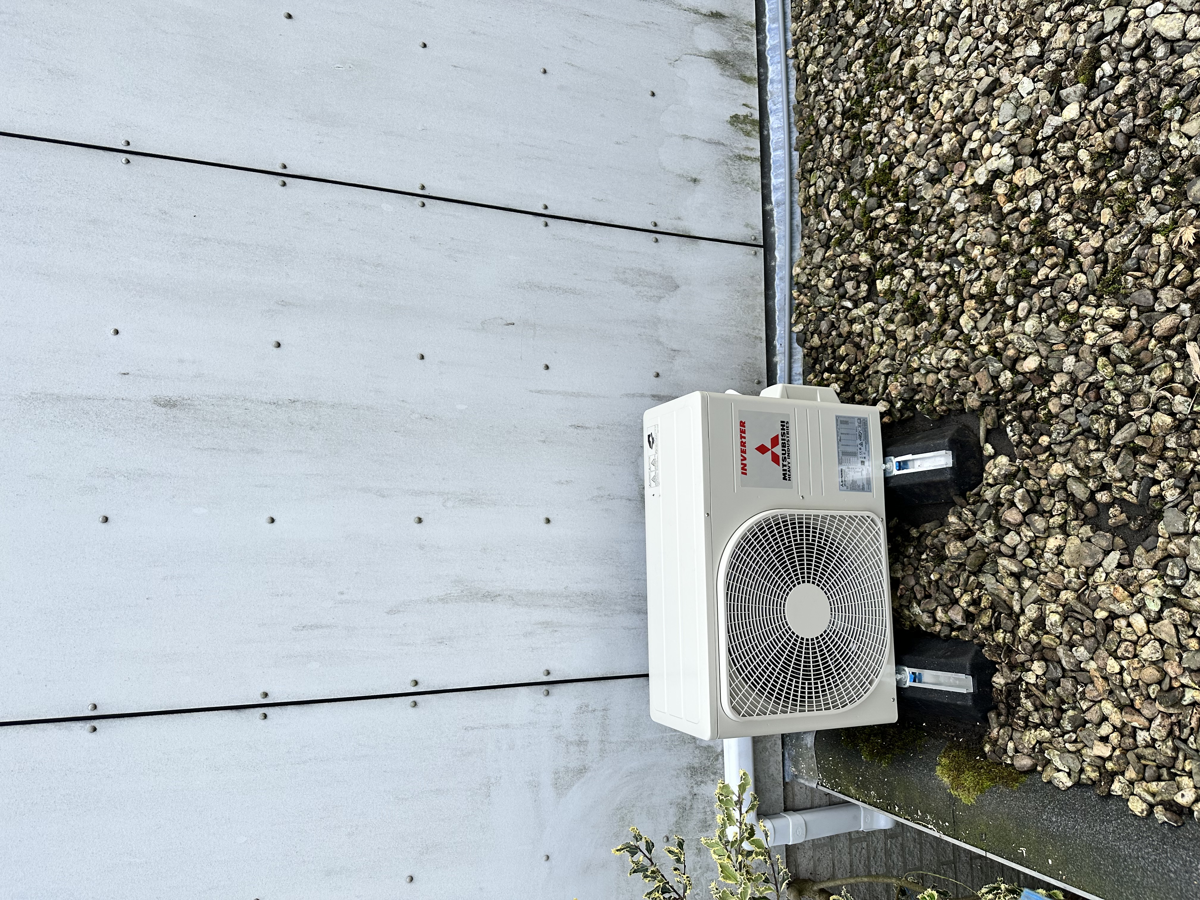 Installatie van een buitenunit van een airconditioningsysteem op een ondergrond van grind, voorzien van een kunststof afvoer.