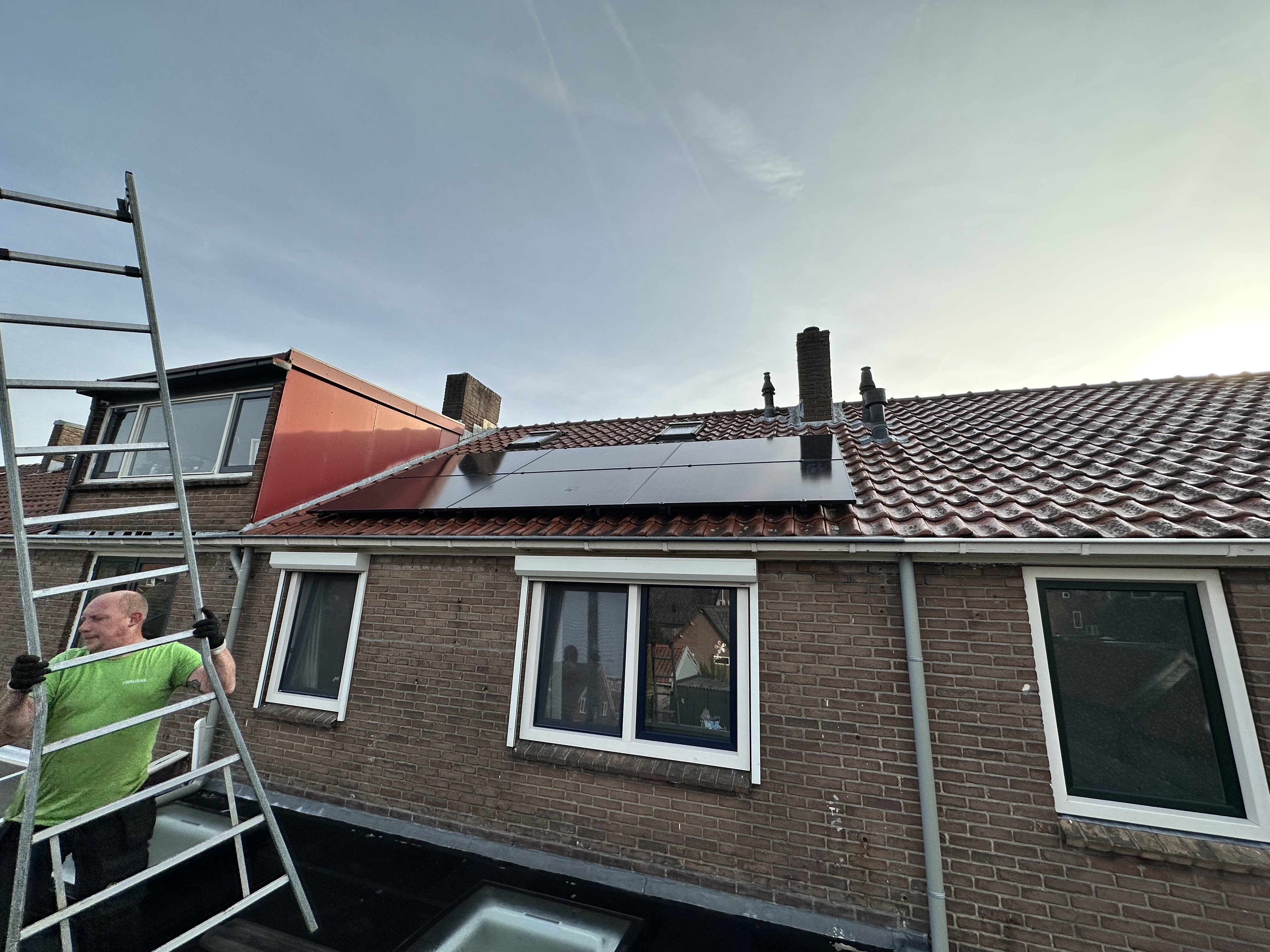 Een installatie van zonnepanelen op een hellend dak met een werknemer die een ladder gebruikt voor de werkzaamheden.