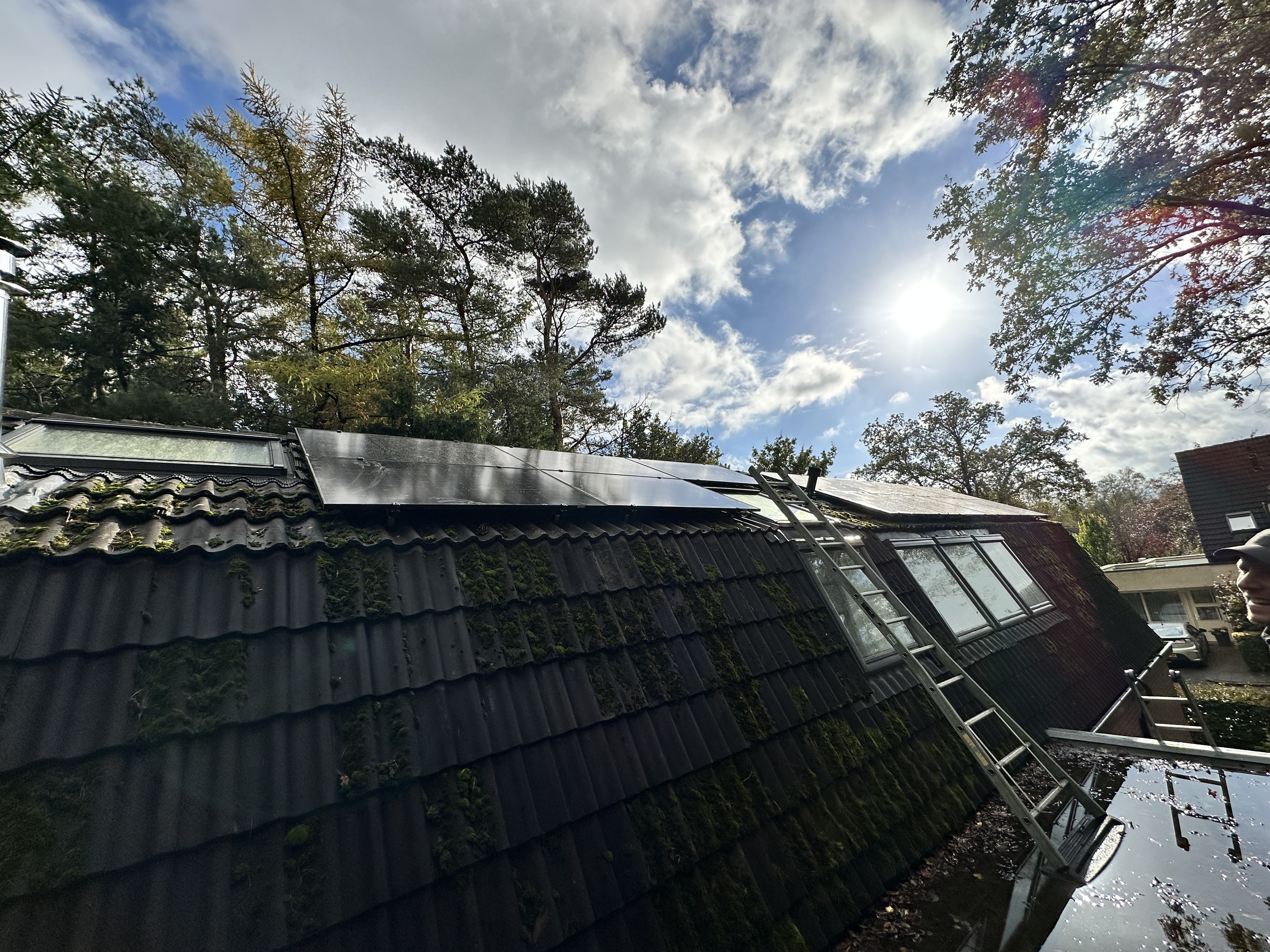 Een installatie van zonnepanelen op een hellend dak, met ladders en bomen op de achtergrond. De panelen vangen zonlicht voor energieproductie.