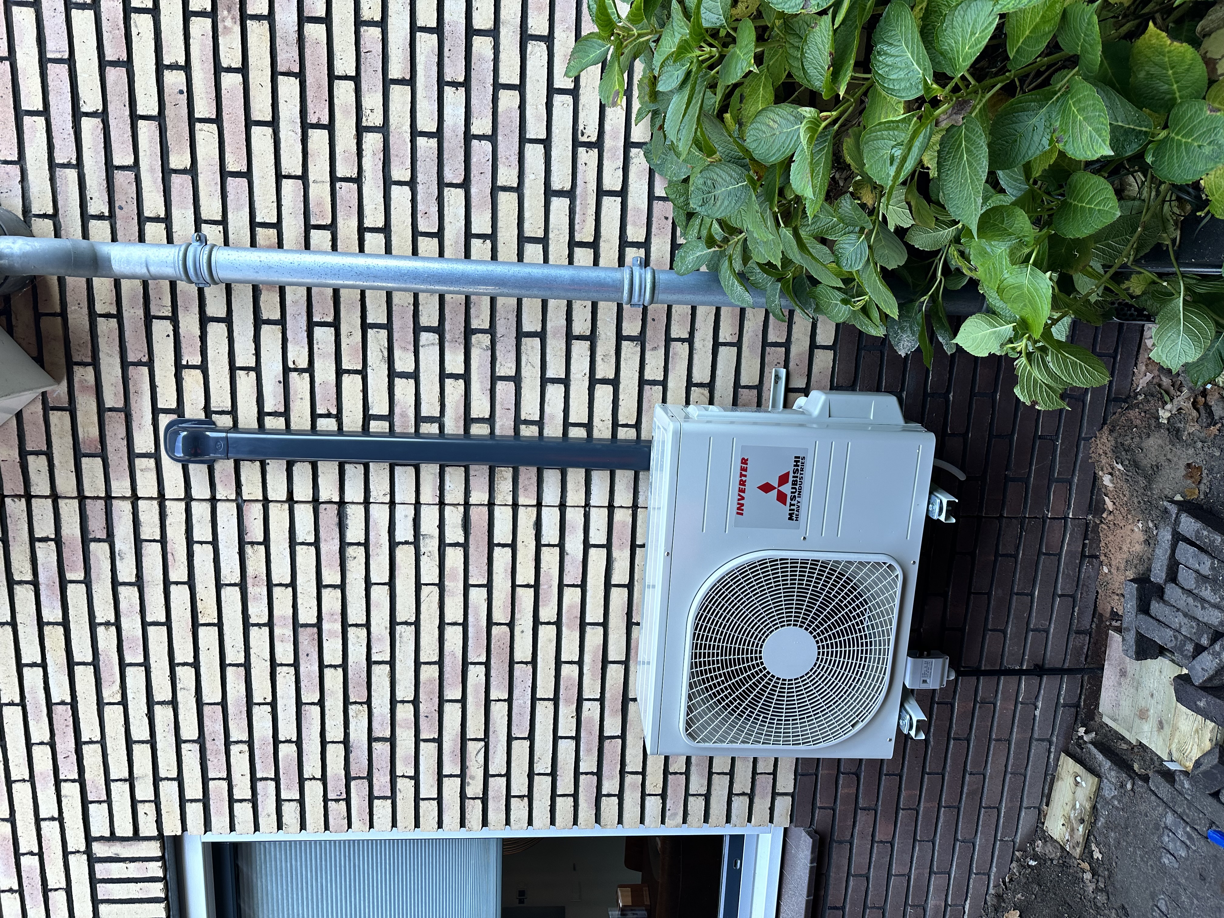 De installatie toont een buitenunit van een airconditioning van Mitsubishi, bevestigd aan een stenen muur met zichtbare leidingen.