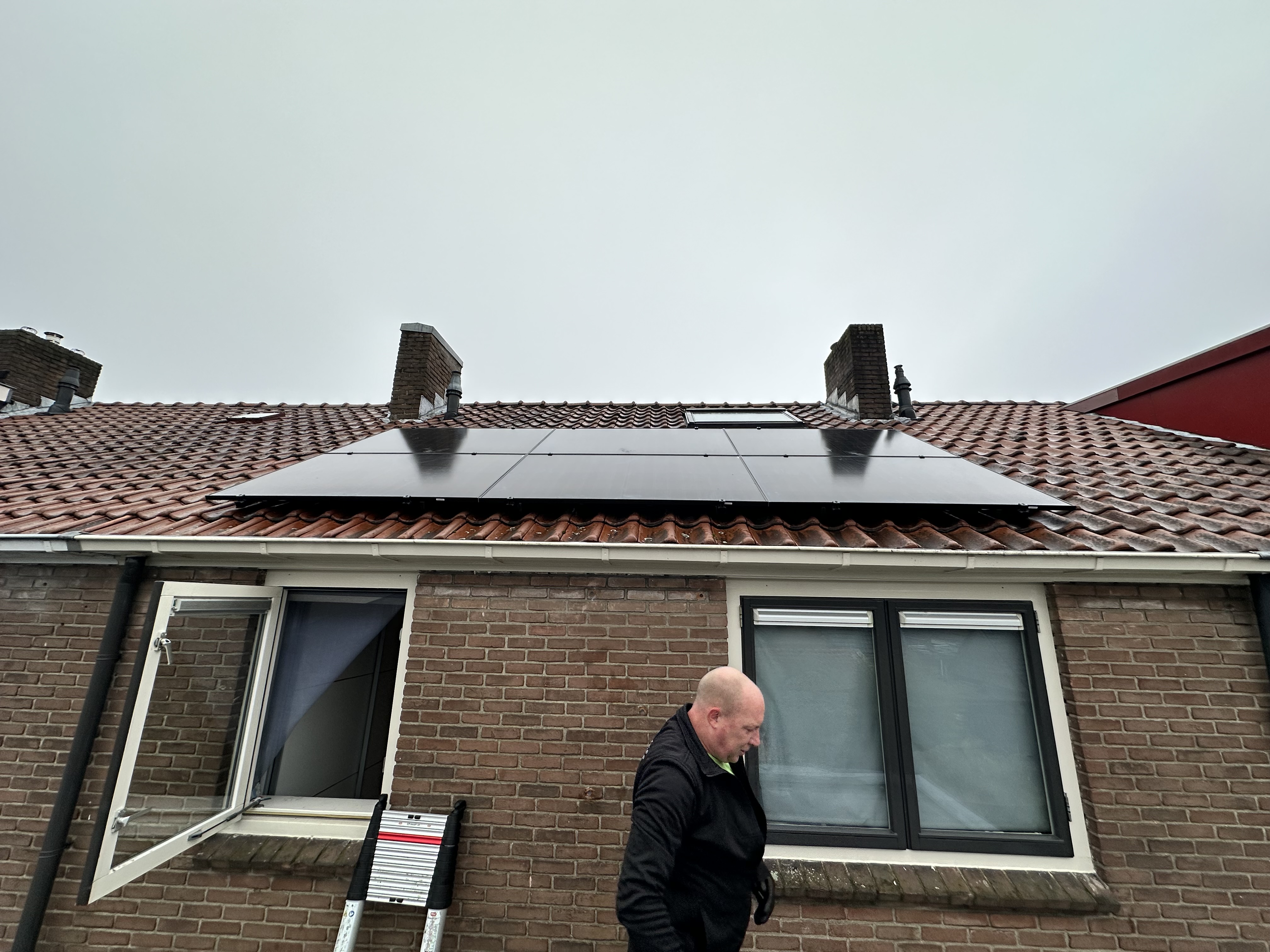 De afbeelding toont een woning met zonnepanelen geïnstalleerd op het schuine dak. Een man loopt ernaast, waarschijnlijk betrokken bij de installatie.