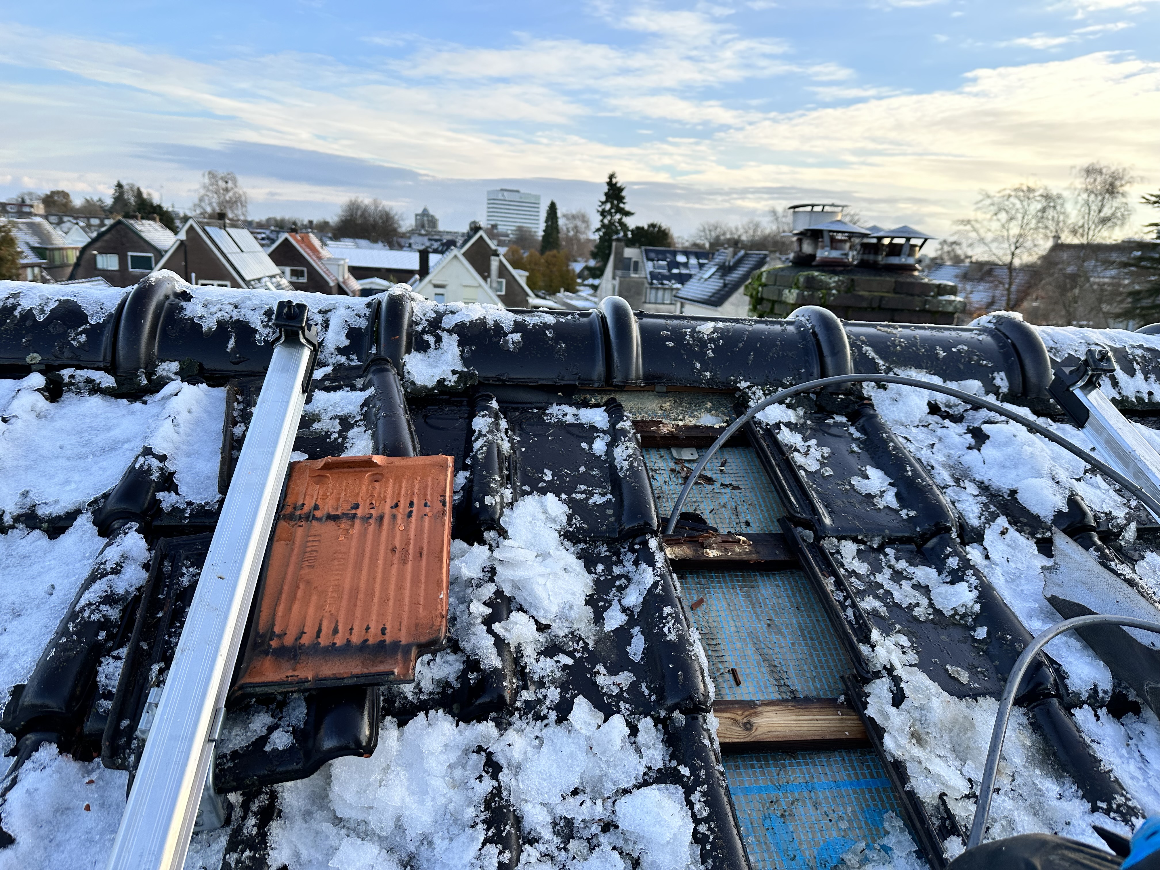 De afbeelding toont een gedeeltelijk geïnstalleerd zonnepaneelsysteem op een dak bedekt met sneeuw, met zichtbare rails en onderdelen.