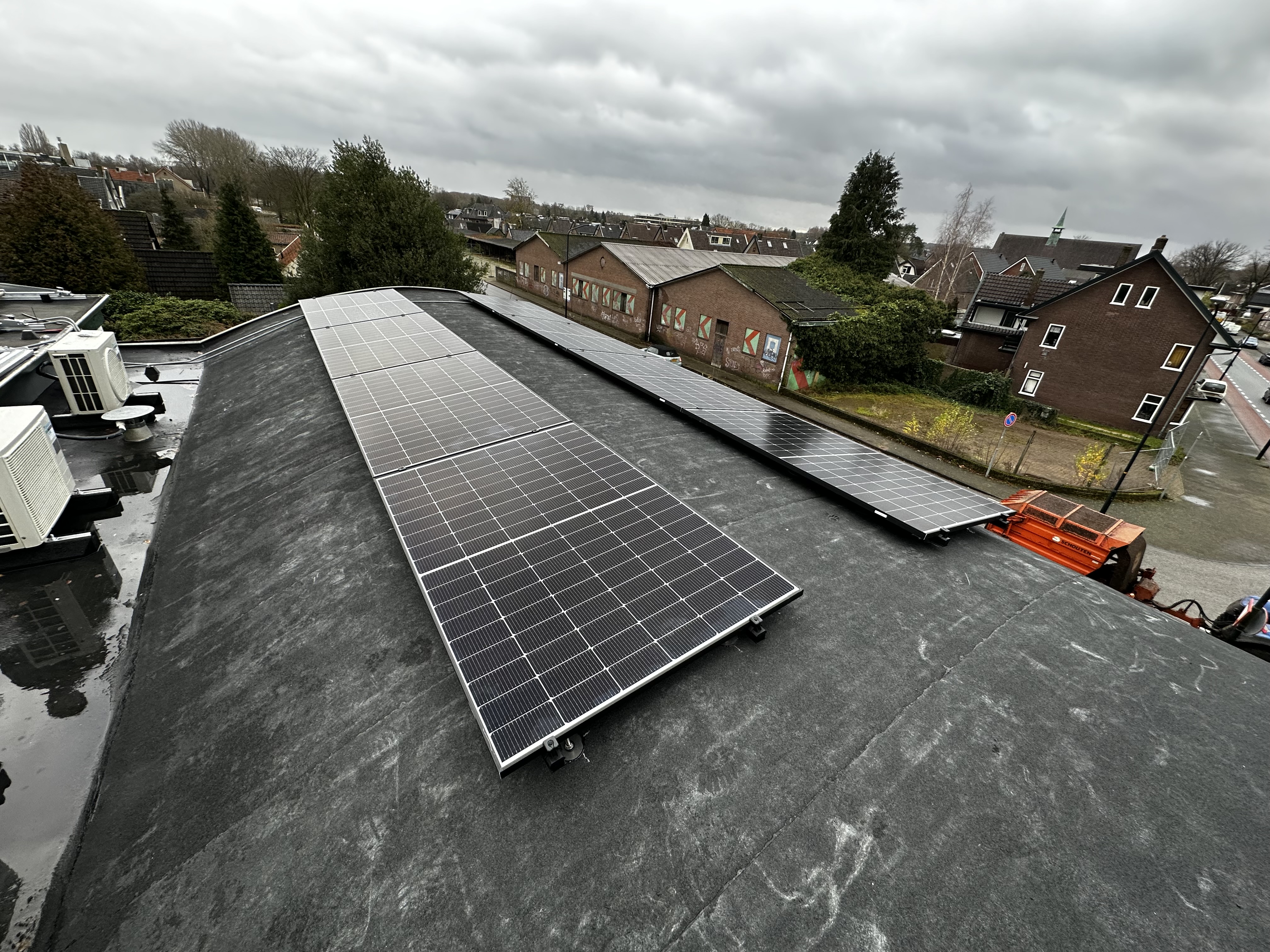De installatie toont een reeks zonnepanelen op een plat dak, met een uitzicht op omliggende gebouwen en vegetatie.