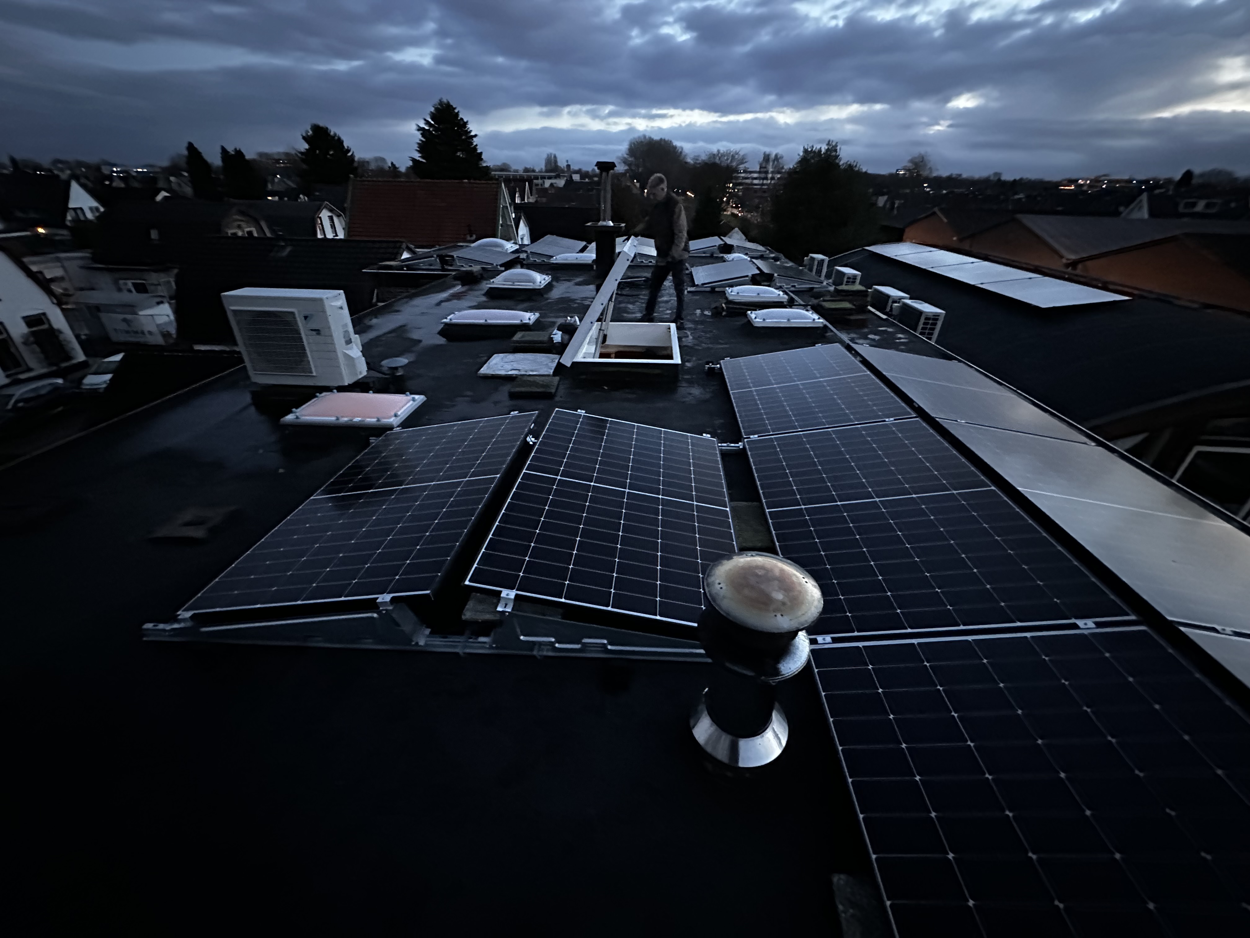 De installatie toont zonnepanelen op een dak, met enkele andere apparaten in de omgeving. De foto is in de schemering genomen.