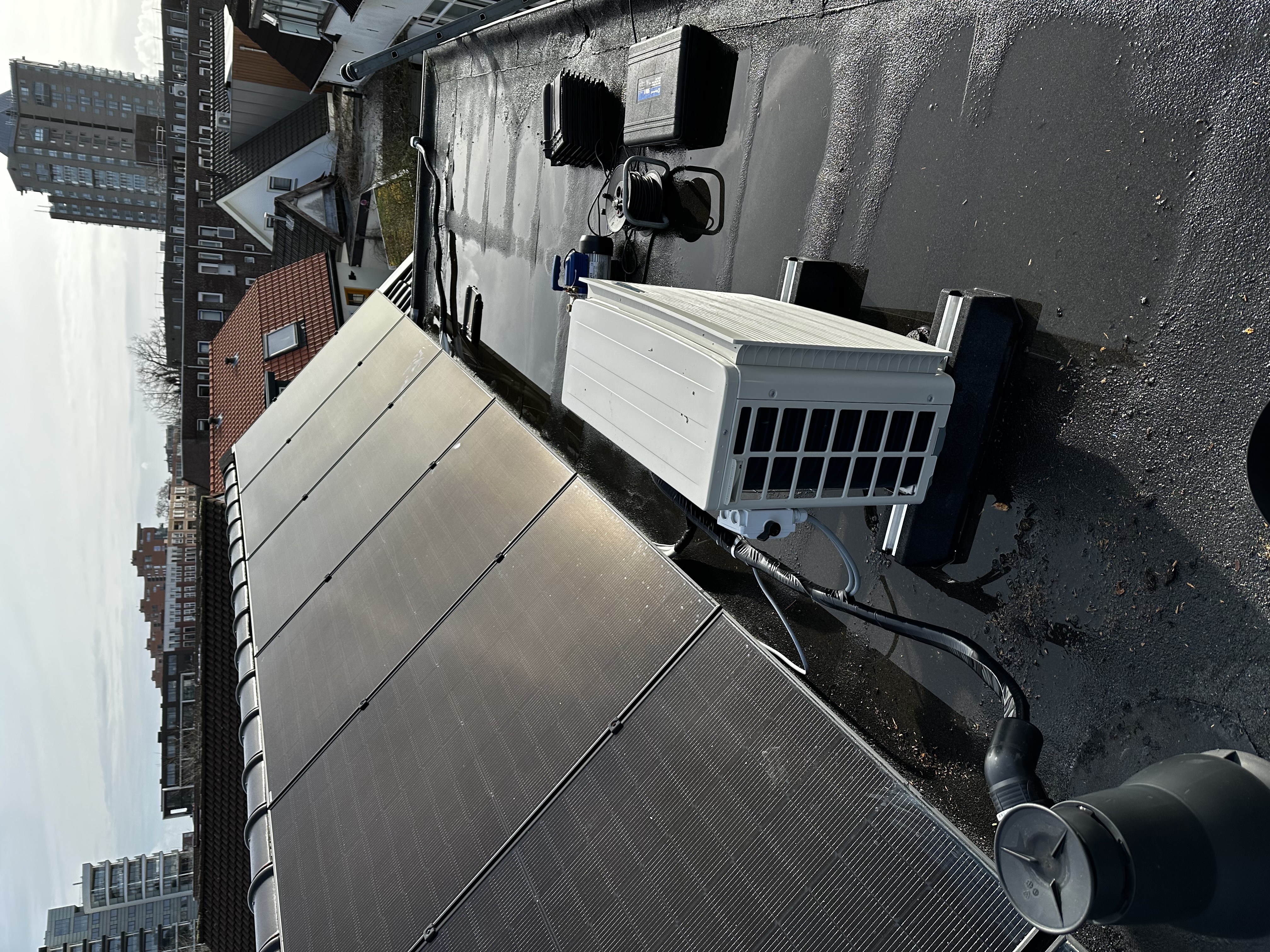 Een airconditioning installatie op een dak, met zonnepanelen op de achtergrond en verschillende aansluitingen zichtbaar.