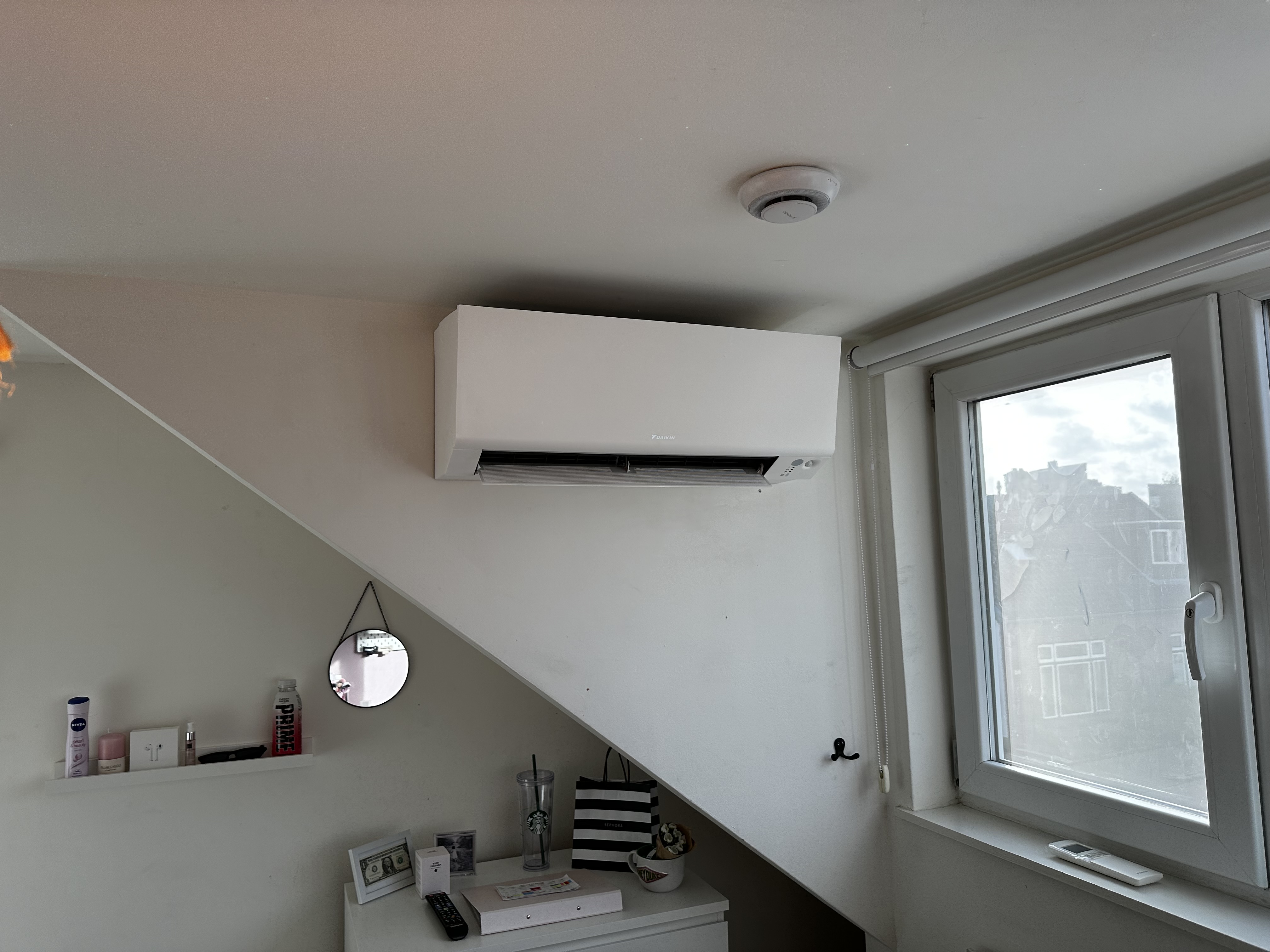 Een wandmontage airconditioner die efficiënt koele lucht verspreidt in een ruimte. Installatie bevindt zich onder een schuin plafond vlakbij een raam.