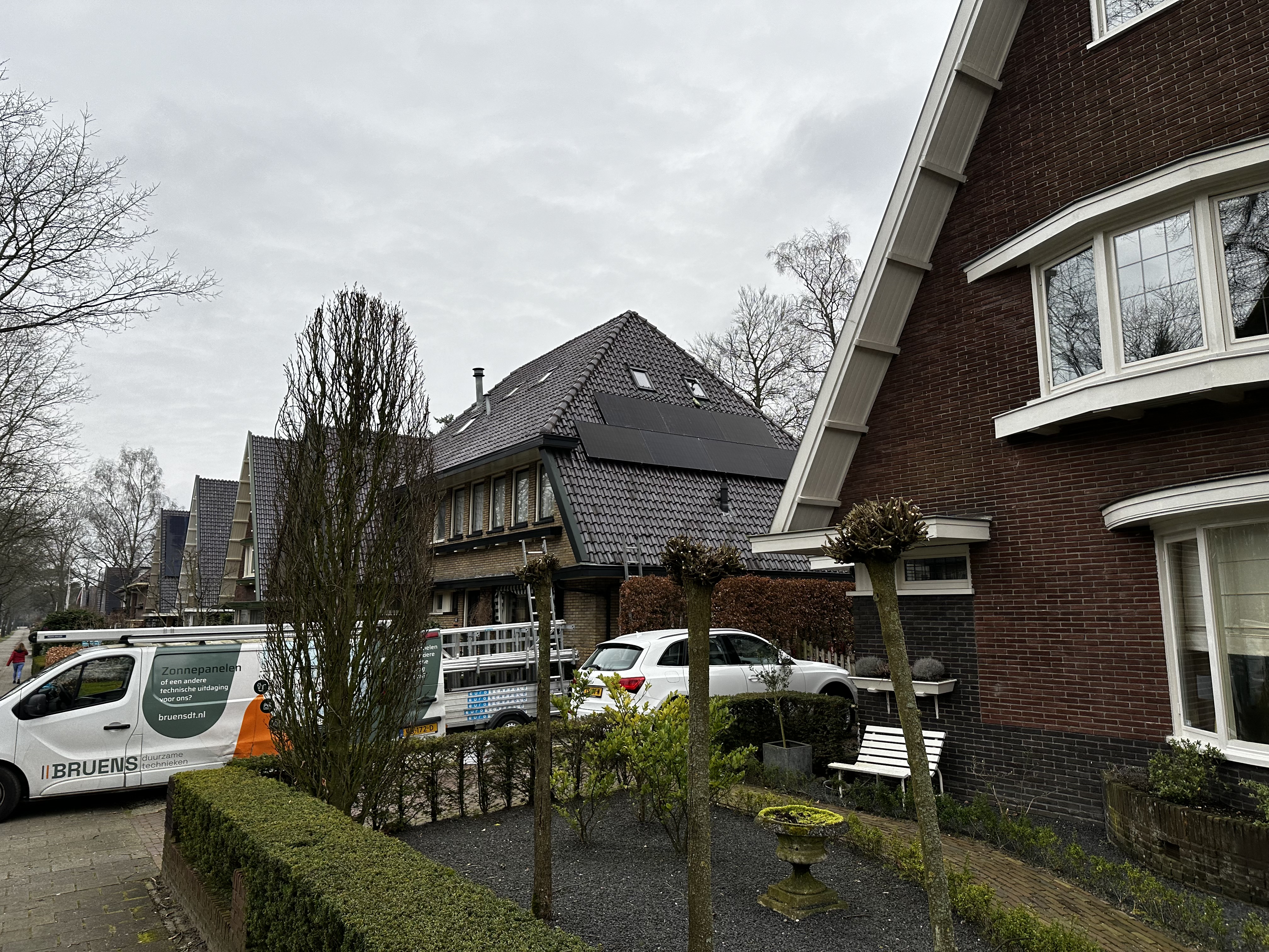 De afbeelding toont een woning met zonnepanelen op het dak, zichtbaar tussen bomen en andere gebouwen.