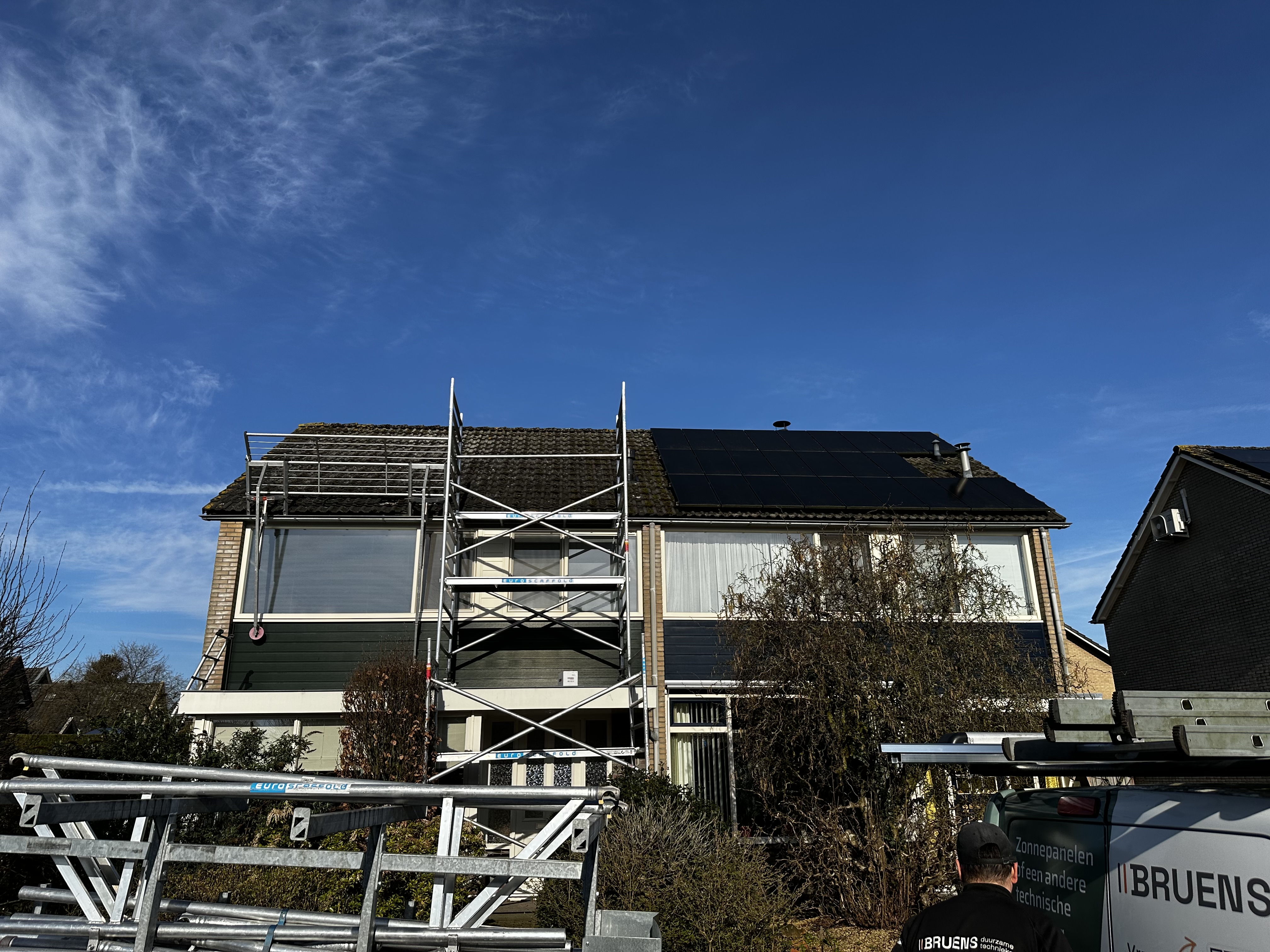 De afbeelding toont een woning met zonnepanelen op het dak, met steigerwerk voor installatie of onderhoud.