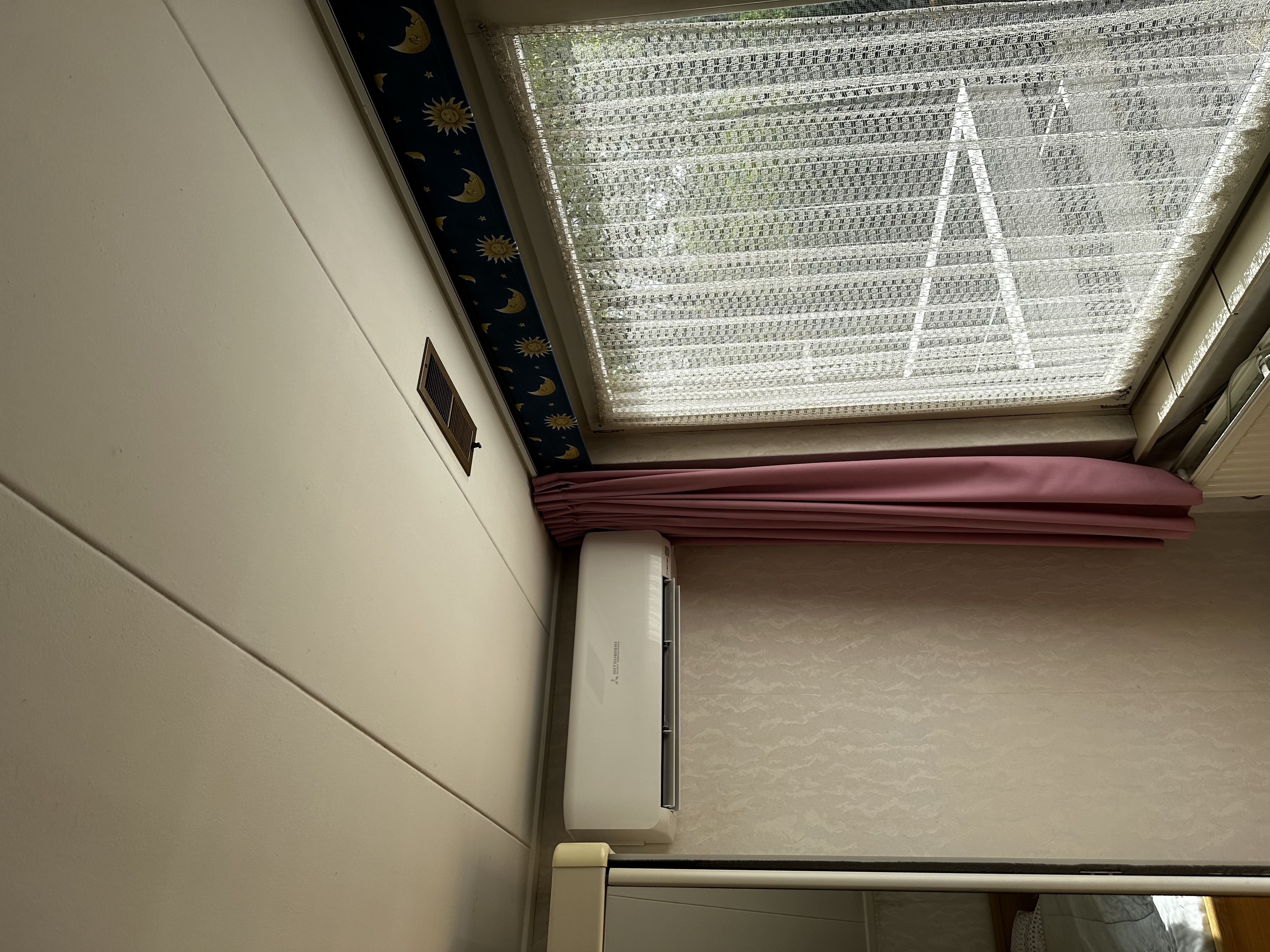 Een wandairconditioner is geïnstalleerd in een kamer, zichtbaar boven een verwarmingselement en een raam met gordijnen.