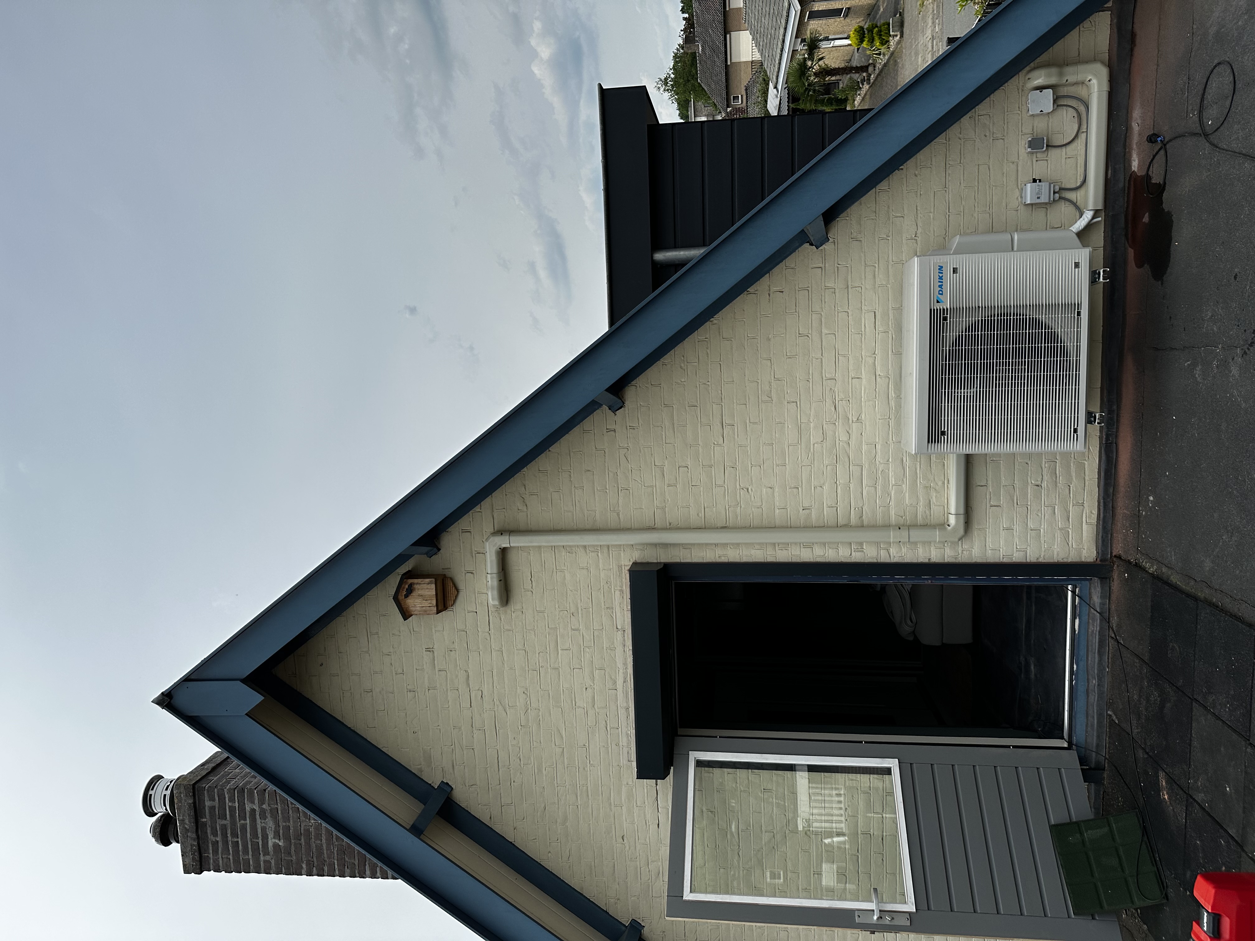 Externe airconditioninginstallatie bevestigd aan de muur van een woning, met leidingen voor koelmiddel en elektriciteit zichtbaar.