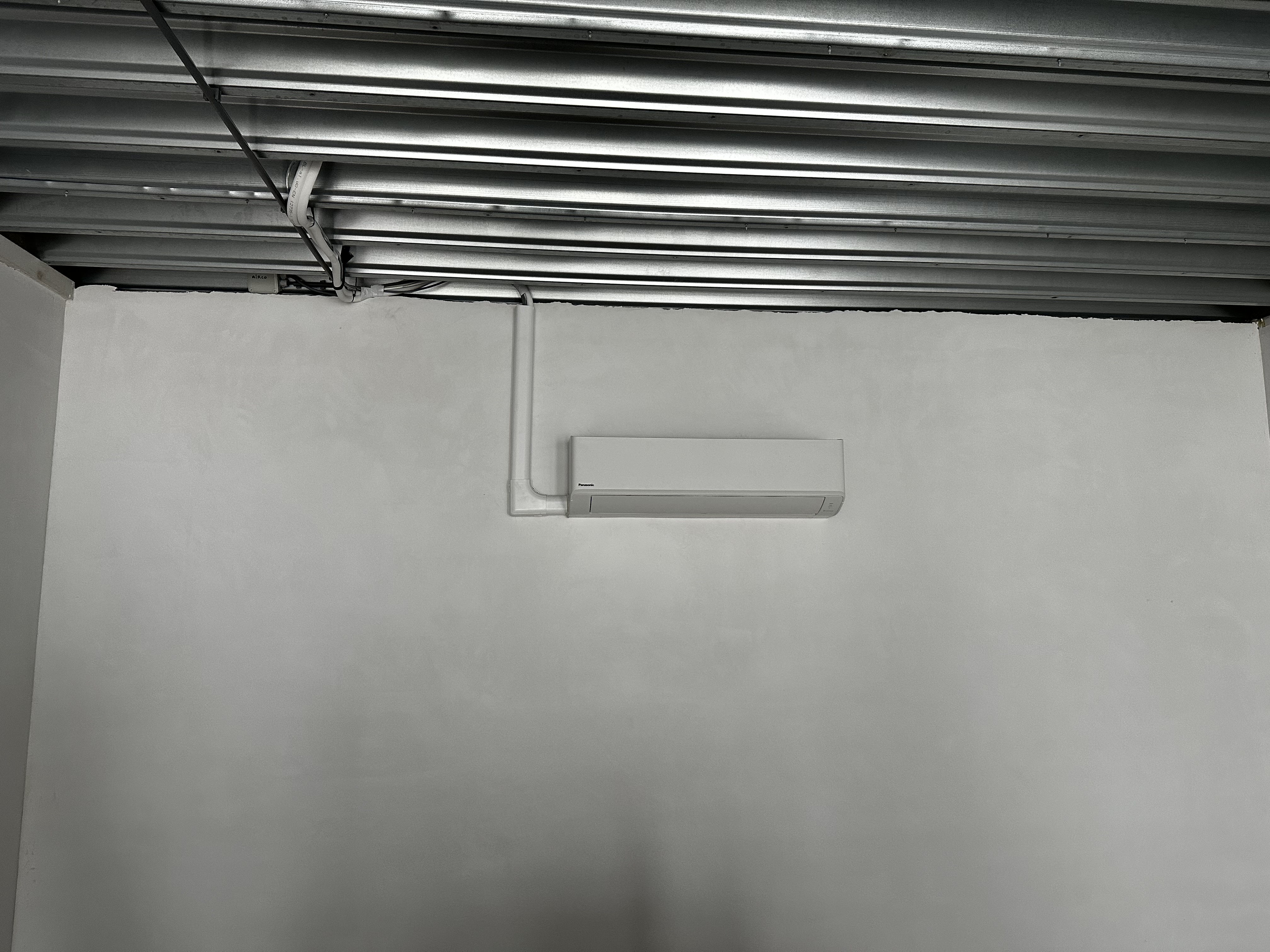 Een wandmodel airconditioningseenheid is geïnstalleerd op een gladde wand, met zichtbare leidingen die de stroomvoorziening en afvoer ondersteunen.