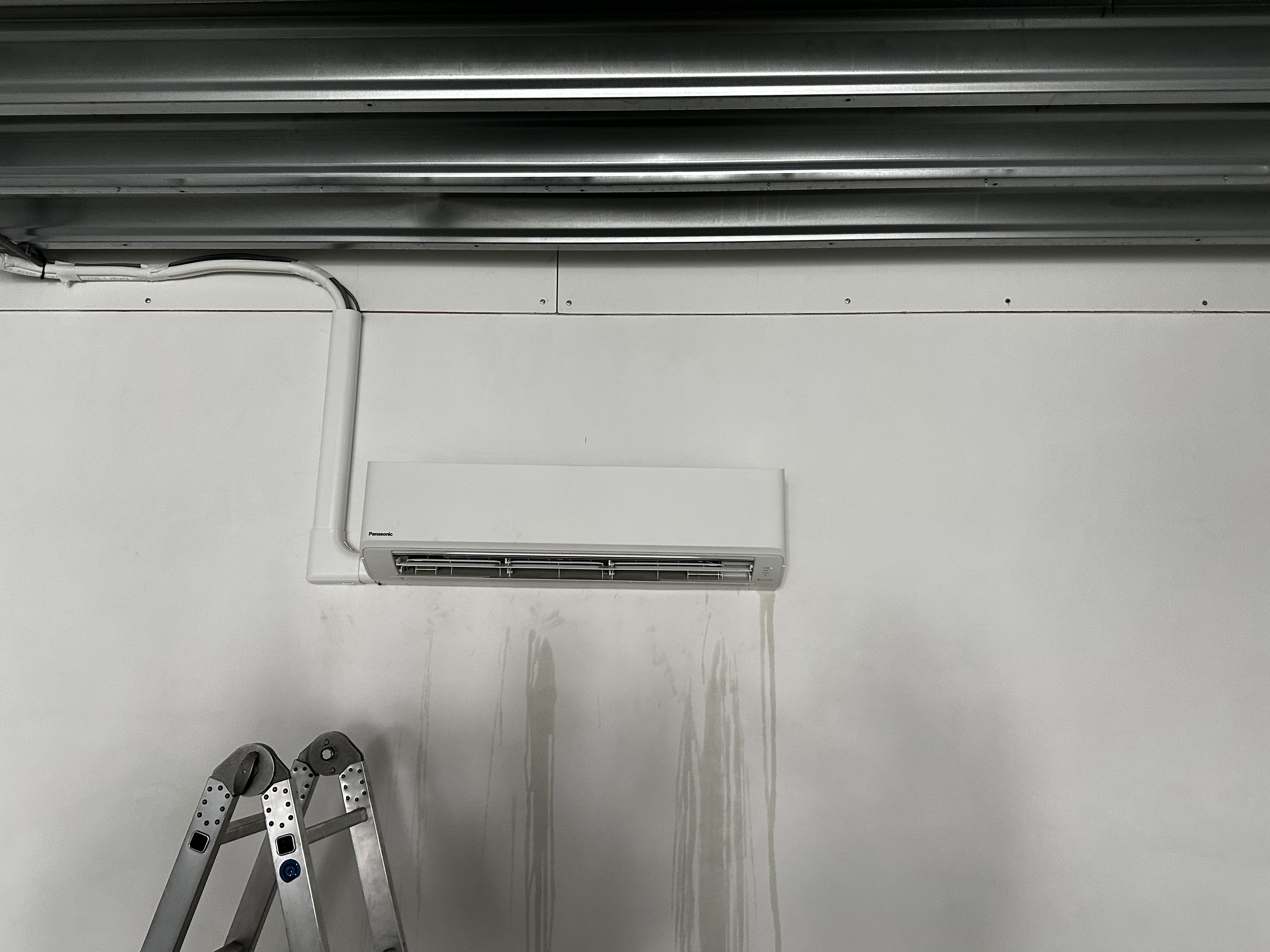 Een wandmontage airconditioner afgesloten met leidingen, zichtbaar op een vlakke muur. Er is een ladder in de hoek zichtbaar, wat wijst op een recent installatieproces.